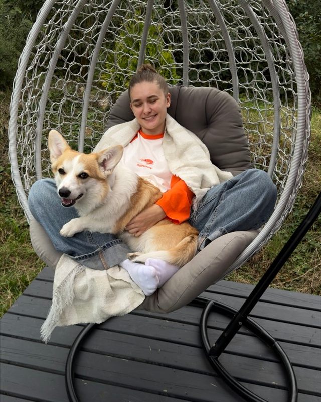 Yuliia smiling in a hanging egg chair with Runa the corgi on her lap