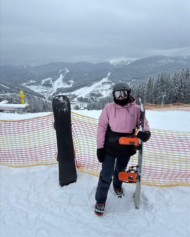 Yuliia in a pink jacket and goggles holding a snowboard on a mountain slope